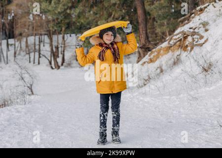 Happy Fun Kind hält einen Plastikschlitten auf dem Hintergrund des verschneiten Waldes. Junge, der Spaß an den weihnachtsferien hat. Konzept des Wintersports, aktive Freizeit Stockfoto