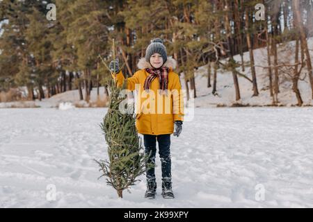 Glückliches Kind hält in den Händen geschnitten weihnachtsbaum auf dem Hintergrund des verschneiten Waldes. Junge mit Weihnachtstanne. Konzept der Winterferien, Traditionen und ce Stockfoto