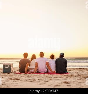 Nehmen Sie sich Zeit für Sonnenuntergänge. Rückansicht einer Gruppe von Freunden, die den Sonnenuntergang am Strand beobachten. Stockfoto