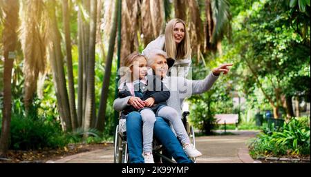 Die Frauen der Familie. Eine fröhliche ältere Frau im Rollstuhl verbringt Zeit mit ihrer Tochter und ihrem Enkelkind draußen in einem Park. Stockfoto
