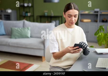 Junges Mädchen mit prothetischem Arm, das ihr Mobiltelefon benutzt, während es am Tisch im Wohnzimmer sitzt Stockfoto