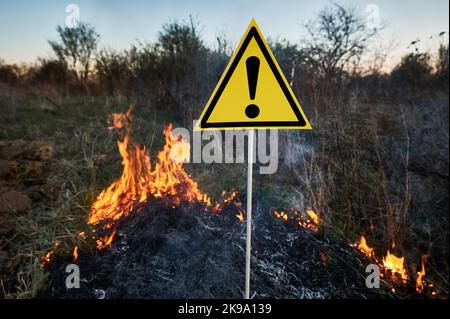 Brennendes trockenes Gras und Warnzeichen mit Ausrufezeichen warnen vor Gefahren im Feld mit Feuer. Konzept von Ökologie und Naturkatastrophen. Stockfoto