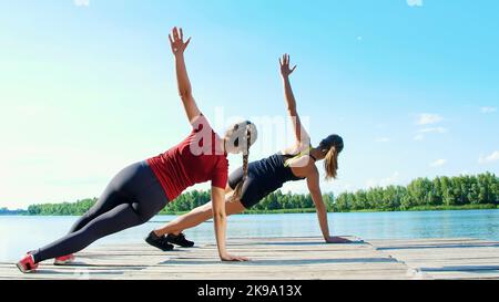 Zwei schöne, athletisch blonde Frauen, die synchron Koordinations- und Gleichgewichtsübungen machen. See, Fluss, blauer Himmel und Wald im Hintergrund, sommerlicher sonniger Tag. Hochwertige Fotos Stockfoto