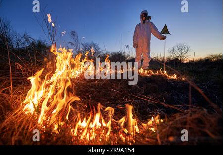 Feuerwehrmann Ökologe arbeitet auf dem Feld mit Wildfeuer in der Nacht. Mann in Anzug und Gasmaske in der Nähe brennendes Gras mit Rauch, mit Warnschild mit Schädel und Kreuzknochen. Konzept für Naturkatastrophen. Stockfoto