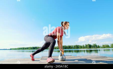 Schöne, athletische junge blonde Frau, die verschiedene Übungen mit Gewichten, Hanteln, Ausfallschritten, Kniebeugen macht. See, Fluss, blauer Himmel und Wald im Hintergrund, sommerlicher sonniger Tag. Hochwertige Fotos Stockfoto