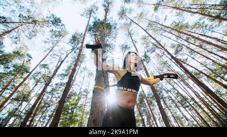 Athletische Frau, Trainerin, Ausbilderin, führt durch, macht Übungen. Im Kiefernwald, im Sommer, in den Sonnenstrahlen. Ansicht von unten, Ansicht von unten. Hochwertige Fotos Stockfoto