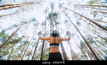 Athletische Frau, Trainerin, Ausbilderin, führt durch, macht Übungen. Im Kiefernwald, im Sommer, in den Sonnenstrahlen. Ansicht von unten, Ansicht von unten. Hochwertige Fotos Stockfoto