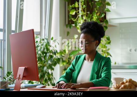 Erfolgreiche afroamerikanische Frau, die als Redakteurin Zeitschrift Artikel tippt, sitzt am Computertisch Stockfoto