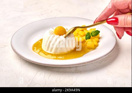 Die Hand der Frau hält einen Löffel über hausgemachter Panna Cotta mit natürlicher Vanilleschote, Mango-Sirup und Minze, selektiver Konzentration Stockfoto