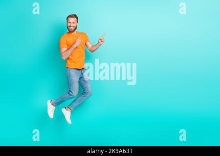 Foto in voller Größe von angenehm schönen Mann muskulösen Bart tragen orange T-Shirt Jeans Regie leeren Raum isoliert auf blauem Hintergrund Farbe Stockfoto