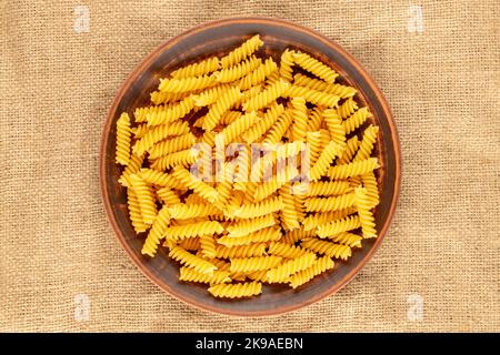 Hartweizen-Pasta in einer Tonplatte auf einem Jutetuch, Makro, Draufsicht. Stockfoto