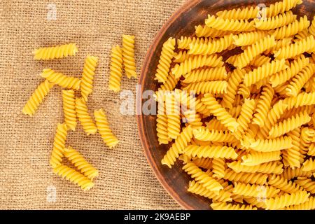 Hartweizen-Pasta in einer Tonplatte auf einem Jutetuch, Makro, Draufsicht. Stockfoto