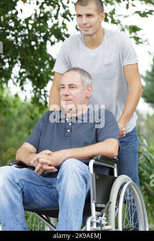 Sohn, der mit einem behinderten Vater im Rollstuhl im Park läuft Stockfoto