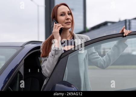 Eine rothaarige, Erwachsene Geschäftsfrau, die auf dem Handy spricht und auf dem Parkplatz aus dem Auto kommt, um ein erfolgreiches Geschäftskonzept zu führen Stockfoto