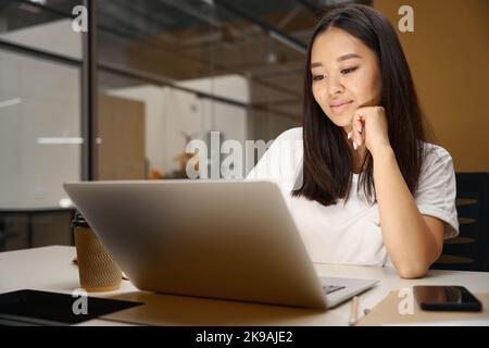 Glückliche asiatische Frau, die im modernen Büro etwas auf einem Notebook sieht Stockfoto