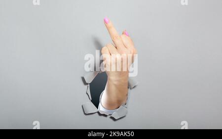 Frau Hand zeigt mit einer Geste, dass alles Mittelfinger . Gerissenes Loch in grauem Papier. Stockfoto