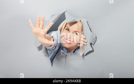 Eine attraktive, fröhliche Frau mittleren Alters, die mit der Hand eine Grußgeste schwenkt, zeigt, dass alles in Ordnung ist. Gerissenes Loch in grau Stockfoto