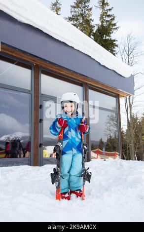 Vorschulkind im Helm, blauer Overall, Skischuhe steht im Schnee, hält Skier in den Händen.Winter aktive Unterhaltung für Kinder, Sportausbildung. Er Stockfoto