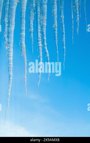 Winterlandschaft mit langen gefrorenen Eiszapfen auf dem Dach auf dem Hintergrund des blauen Himmels, natürliche lange Eiszapfen, Winter Natur Hintergrund Stockfoto