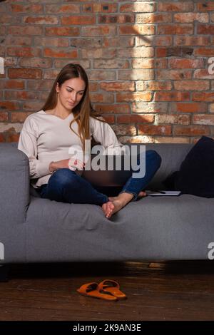 Frau in einem weißen Pullover, die mit einem Laptop arbeitet, mit einem Notebook an der Seite und Schuhen vor ihr Stockfoto