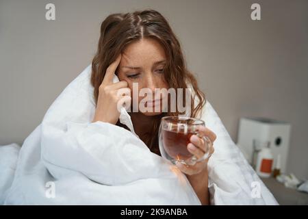 Frau, die im Bett saß und mit einer Decke mit einer Tasse Tee bedeckt war Stockfoto