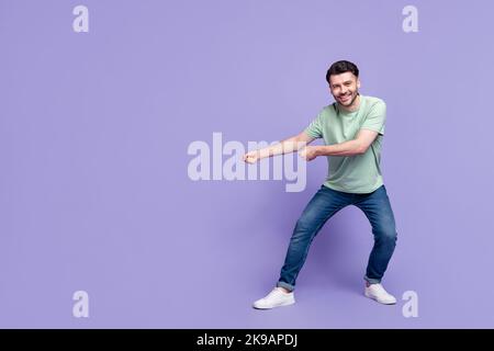 Ganzkörper-Foto von attraktiven jungen Mann ziehen Seil leeren Raum fröhlich gekleidet stilvollen grauen Kleidungsstück auf lila Hintergrund isoliert Stockfoto