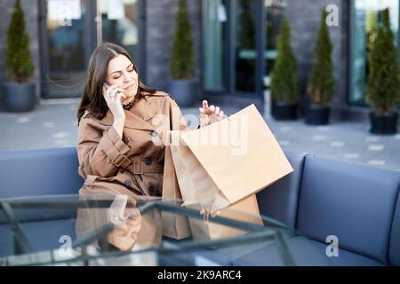 Seitenansicht der jungen Frau mit sitzt auf Holzbank Papiertaschen mit Käufen um nutzt Handy scrollt durch neue Nachrichten Stockfoto