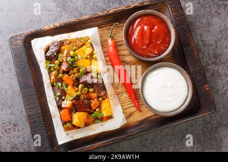 Türkischer Kebab mit Kartoffeln, grünen Erbsen, Zwiebeln und Karotten, in Papier eingewickelt und in der Nähe auf einem Holztablett auf dem Tisch gebacken. Horizontale Ansicht von oben Stockfoto
