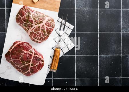 Steaks Raw. Rohes Cowboy-Steak, marmoriertes Rindfleisch mit Gewürzen Rosmarin und Pfeffer auf weißem Marmorschiefer und grauem Tischhintergrund. Draufsicht. Modell. Stockfoto