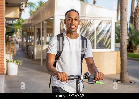 Glücklich lächelnder afroamerikanischer Mann in lässiger Kleidung, der an einem sonnigen Tag einen Elektroroller auf der Straße fährt. Fröhlicher, hübscher Mann, der einen Roller reitet Stockfoto