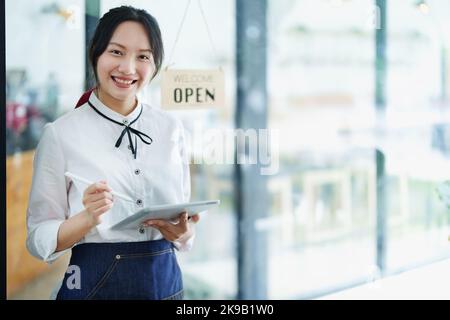 Eine junge asiatische Frau, die ein kleines Unternehmen eröffnet und ein lächelndes Gesicht zeigt, hält ein Tablet in einer Schürze, die vor einer Café-Bar steht Stockfoto