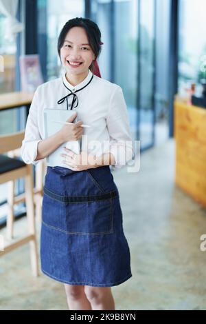 Eine junge asiatische Frau, die ein kleines Unternehmen eröffnet und ein lächelndes Gesicht zeigt, hält ein Tablet in einer Schürze, die vor einer Café-Bar steht Stockfoto