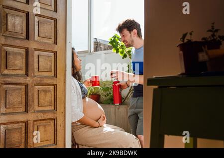 Das junge, schwanger lateinische Paar trinkt heiße Getränke und spricht zu Hause, durch die Eingangstür vom Haus aus gesehen. Stockfoto