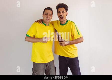 Black Brothers mit gelbem brasilianischem Hemd und Bierflaschenjubeln isoliert auf Weiß. Fußballfans jubeln über den Sieg Brasiliens. Stockfoto