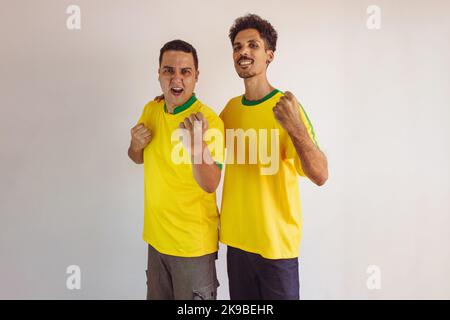 Black Brothers mit gelbem brasilianischem Hemd und Flaggenjubel isoliert auf Weiß. Fußballfans jubeln über den Sieg Brasiliens. Stockfoto