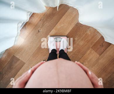 Draufsicht auf die werdende Mutter, die zum Wiegen auf der Bodenwaage steht und ihre beiden Handflächen auf den Bauch legt. Eine Schwangerin misst ihr Körpergewicht zu Hause mit einer Personenwaage. Stockfoto