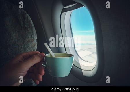 Die Hand eines Mannes hält eine Tasse Kaffee vor dem Flugzeugfenster, in dem Sie den blauen Himmel und den Flügel sehen können Stockfoto