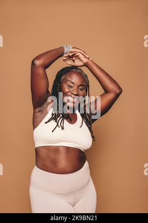 Glückliche Frau lächelt vor der Kamera, während sie vor einem braunen Hintergrund steht. Sportliche Frau mit Dreadlocks in Sportkleidung in einem Studio. Reif Stockfoto