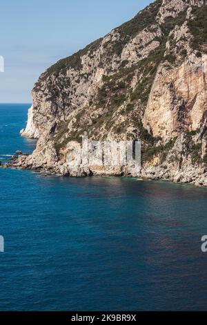 Korfu, Griechenland. Ein riesiger Bergfelsen, auf dem Grün wächst, wächst in der Nähe des schönen blauen Wassers, wo die Sommersonne scheint. Stockfoto