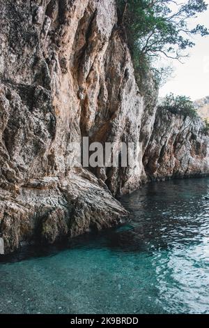 Korfu, Griechenland. Ein riesiger Felsen am Strand mit blauem Wasser, auf dem grüne Pflanzen und Bäume wachsen. Stockfoto