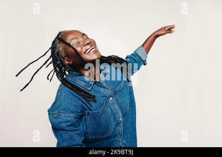 Glück im Studio. Reife Frau mit Dreadlocks, die mit geschlossenen Augen und ausgestreckten Armen lacht. Fröhliche Frau mittleren Alters, die einen deni trägt Stockfoto