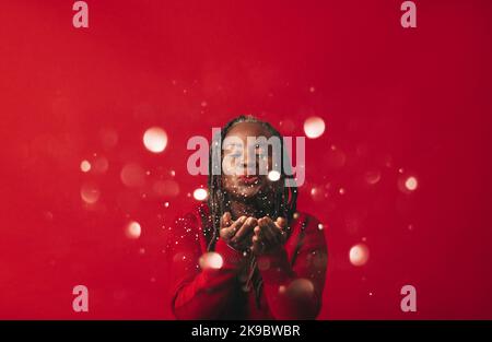 Glückliche schwarze Frau, die Konfetti in die Luft bläst, während sie vor einem roten Hintergrund steht. Reife Frau mit Dreadlocks feiern und Spaß in einem Stockfoto