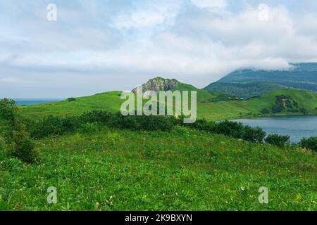 Natürliche Landschaft der Insel Kunashir mit grasbewachsenen Hügeln, vulkanischen Felsen, Vulkan in den Wolken, Lagune und Ozean in der Ferne Stockfoto