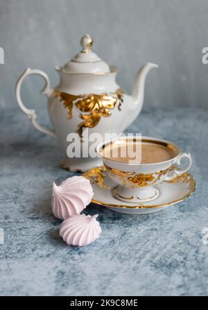 Edle weiße und vergoldete Kaffeetasse auf einem abstrakten blauen Hintergrund. Kaffee mit einem appetitlich goldenen Schaum. Zwei rosa lockige Baiseumen neben der Tasse. B Stockfoto