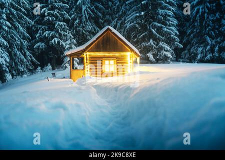 Fantastische Winterlandschaft mit glühender Holzhütte im verschneiten Wald. Gemütliches Haus in den Karpaten. Weihnachtsfeiertagskonzept Stockfoto