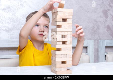 Interessiert glücklich kleinen blonden Jungen spielen Brettspiel unter Ziegel aus Holzturm Gleichgewicht halten. Brettspiel Turm aus Holzblöcken. Aktivität für Stockfoto