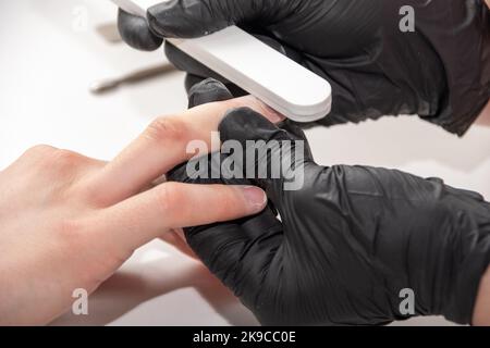 Handschuhen eines erfahrenen Manikuristen, der die Nägel einer jungen Frau mit einer Nagelfeile feilte. Hände während einer Maniküre-Pflege in einem Spa-Salon. Manikurist Fi Stockfoto