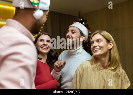 Vier verschiedene Freunde feiern und tanzen auf der Silvesterparty, die Hausgäste amüsieren sich an Weihnachten, in festlicher Neujahrskleidung und Weihnachtsmützen im Wohnzimmer. Stockfoto