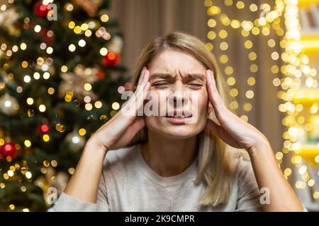 Eine junge, schöne Frau sitzt zu Hause, hält ihren Kopf mit den Händen und grimazt vor Schmerzen. Sie fühlt starke Kopfschmerzen. Sie ist es leid, sich auf das neue Jahr und die Weihnachtsfeiertage vorzubereiten. Stockfoto