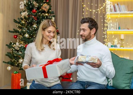 Ein verliebter Mann und eine verliebte Frau an Weihnachten feiern Neujahrsferien und tauschen Geschenke aus, die zu Hause auf dem Sofa in der Nähe des Weihnachtsbaums im Wohnzimmer sitzen. Stockfoto